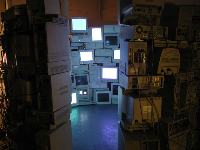A large, cylindrical structure made entirely of CRT monitors and towers facing inwards. There is a gap where someone could walk in. Some of the screens display very light cool colors.